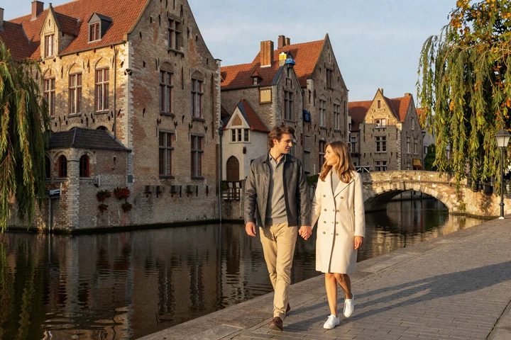 Couple walking hand in hand along a picturesque Bruges canal at golden hour with medieval buildings